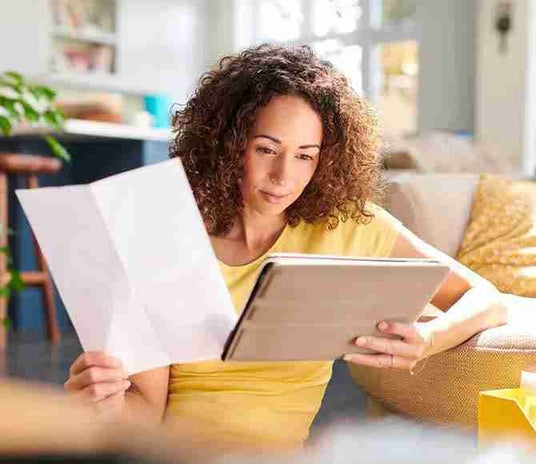 Woman reviewing appliance options on a tablet while holding papers in a bright living room.